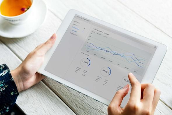 A person holding a tablet displaying a finance report with line graphs and circular charts, sitting at a white wooden table next to a cup of tea.