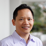 A man with short black hair smiles while wearing a light purple collared shirt. He is indoors with a blurred background of greenery and white walls.