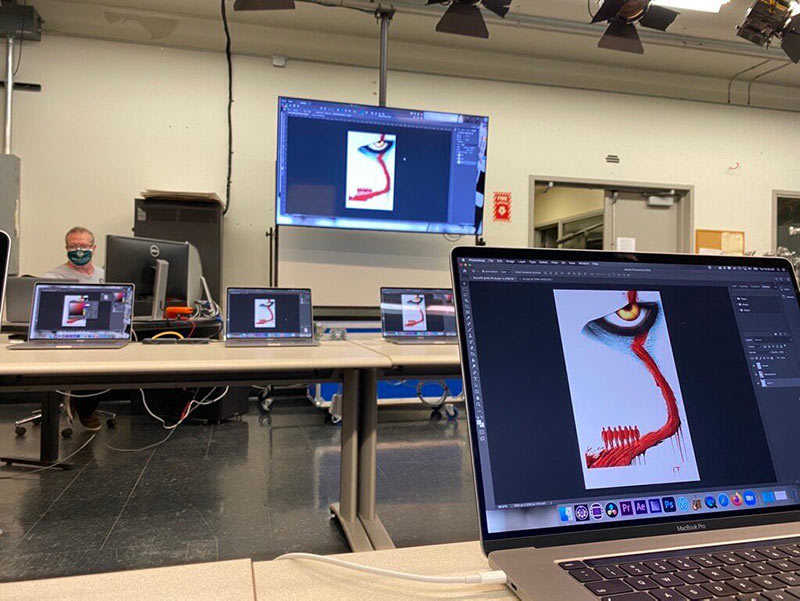 A classroom with several laptops and monitors displaying digital artwork of a stylized eye and red lines. A person wearing a mask sits at a desk in the background. The room has overhead lights and neutral-colored walls.