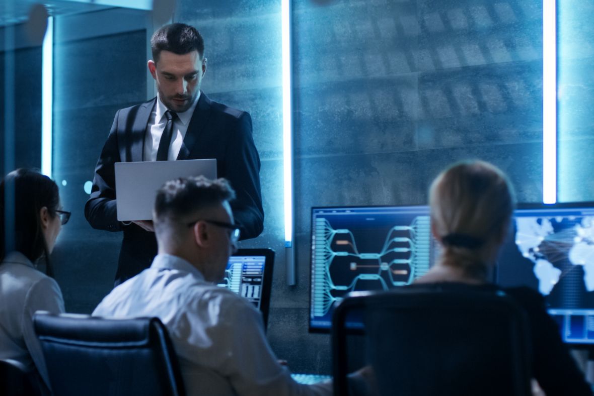 A man in a suit holds a laptop while standing behind three people working at computer monitors, displaying data and world maps, in a dimly lit, modern office or control room.