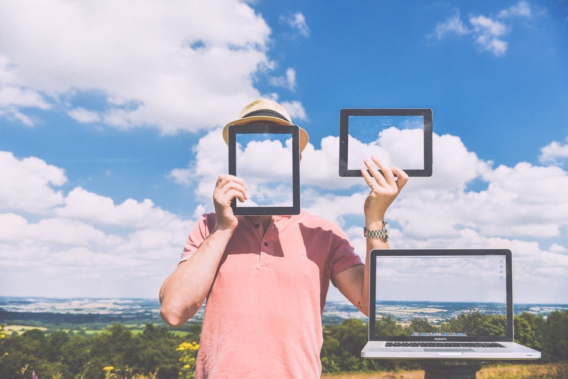 A person in a pink shirt and hat holds a tablet and a smartphone that display the sky, blending with the background. A laptop showing the same sky is on their side, creating an illusion in a scenic outdoor setting.