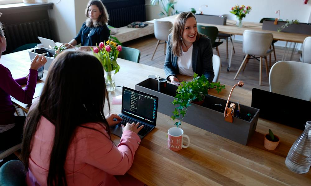 A group of women sitting at a conference desk in the office on their laptops.