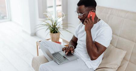A man sitting on a couch with a laptop and talking on a smartphone while working remotely.