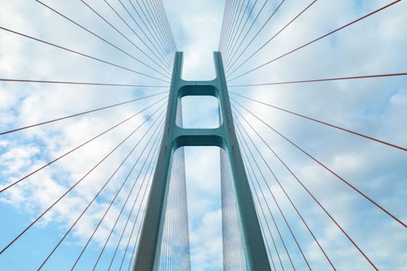 View looking up at a modern cable-stayed bridge against a partly cloudy blue sky, with numerous cables radiating from the central tower structure.