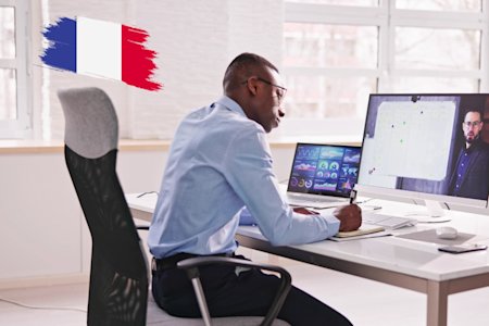 A man in a blue shirt sits at a desk, working on a computer with graphs on the screen while participating in a video call. A French flag graphic is visible on the wall beside him.