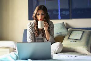 A woman sitting on her bed and drinking a coffee while using a laptop to access a remote desktop.