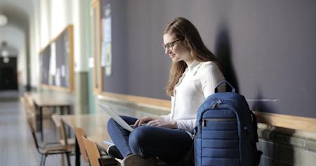 A female student sitting on a desk with her laptop in her lap and a backback next to her.