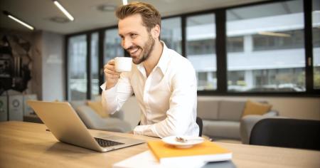 A smiling man sipping coffee while working at his laptop.