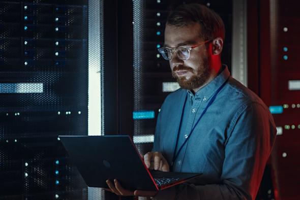 A man with glasses and a beard is using a laptop while standing in a server room with illuminated server racks in the background. The lighting is blue and red, creating a tech-focused atmosphere.
