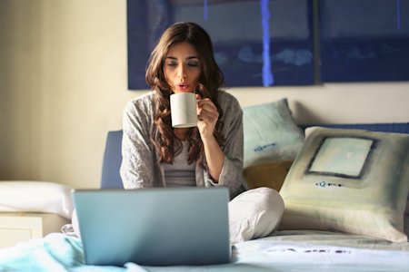 A woman sitting on her bed working on a laptop an drinking coffee.