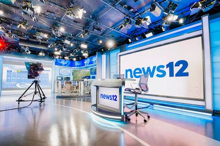 A brightly lit TV news studio with a large screen displaying news12, a news desk, chairs, cameras, and monitors, featuring a modern blue and white design.