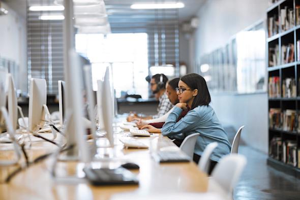 Several people work at desktop computers in a bright, modern library or computer lab. Bookshelves line the wall, and large windows let in natural light. The focus is on a woman in glasses concentrating on her screen.