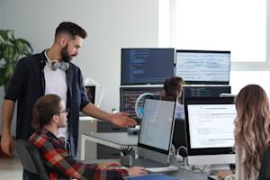 Four people work together in a modern office with multiple computer monitors displaying code; one man stands and gestures at a colleague’s screen, while others sit and focus on their tasks.