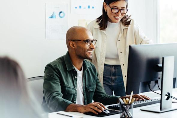 Two people are smiling and working together at a computer in an office. One is seated and typing, while the other stands beside him, looking at the monitor. Charts are visible on the wall in the background.