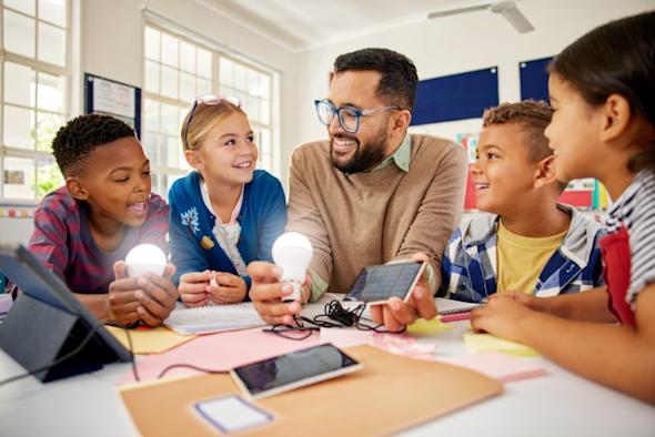 A teacher and four smiling children sit around a table in a classroom, holding glowing light bulbs and a smartphone, engaging in a hands-on science activity. Notebooks, tablets, and papers are on the table.