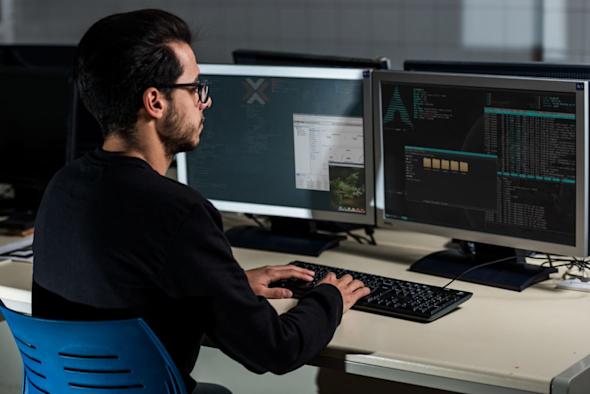 A person with dark hair and glasses works at a desk with dual monitors displaying code and system information in a dimly lit office environment.