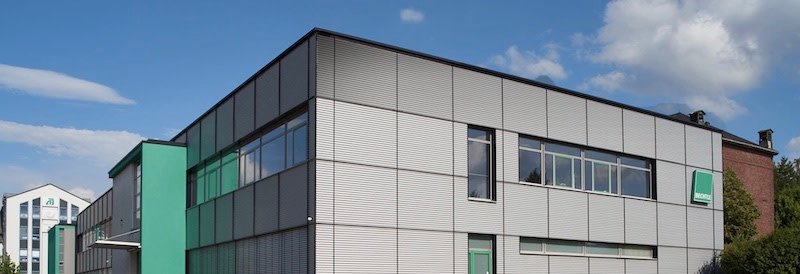 A modern, rectangular industrial building with gray siding and green accents, large windows, and a clear blue sky with some clouds in the background.