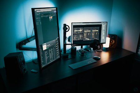 A modern desk setup with a vertical monitor displaying code, a horizontal monitor, keyboard, mouse, speakers, headphones, a lamp, and various desk accessories, all illuminated by blue-toned lighting.
