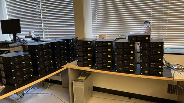 A room with two desks holding numerous stacked desktop computer towers. Some monitors, cables, and office supplies are visible; horizontal blinds cover the windows in the background.