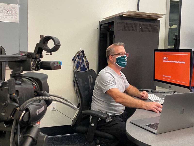 A man wearing a face mask sits at a desk, working on a computer. A camera is set up nearby, and the computer screen displays Lab 2: Getting Started with Adobe Photoshop in a classroom or lab setting.