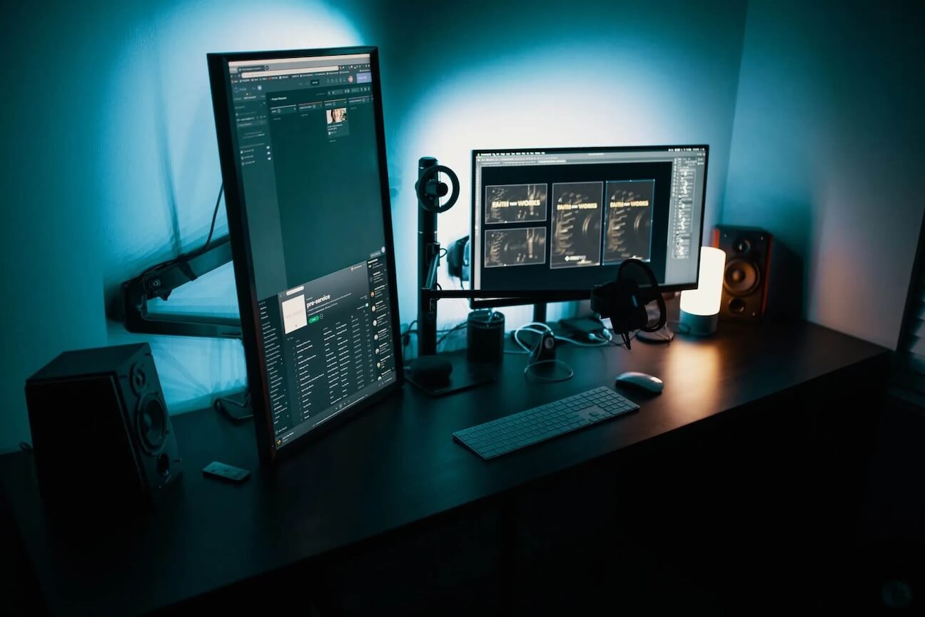A modern desk setup with a vertical monitor displaying code, a horizontal monitor, speakers, a keyboard, headphones, a microphone, a lamp, and ambient blue lighting in a dim room.