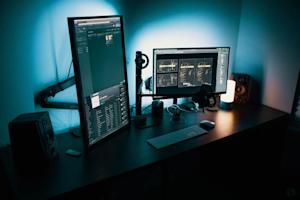 A modern desk setup with a vertical monitor displaying code, a horizontal monitor, speakers, a keyboard, headphones, a microphone, a lamp, and ambient blue lighting in a dim room.