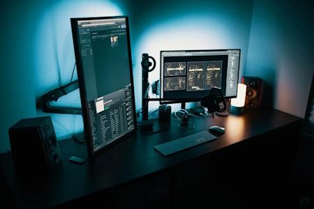 A modern desk setup with a vertical monitor displaying code, a horizontal monitor, speakers, a keyboard, headphones, a microphone, a lamp, and ambient blue lighting in a dim room.