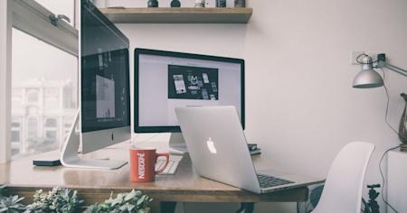 A Mac laptop on a desk in front of two other Apple monitors.