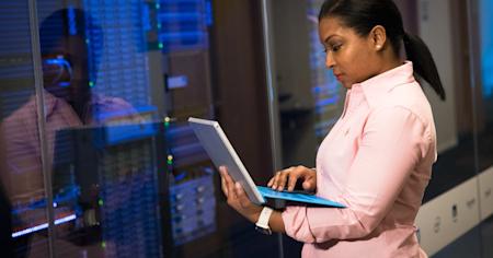 A woman standing in a server room while working on her laptop.