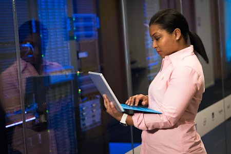 A woman standing in a server room while working on her laptop.