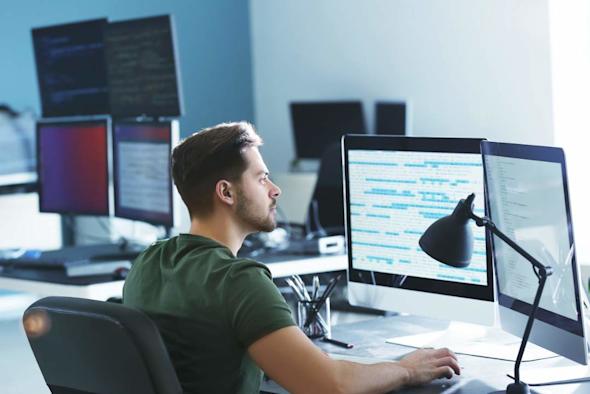 A man sits at a desk in an office, working on a computer with multiple monitors displaying code. A desk lamp, pens, and other office supplies are nearby. Sunlight streams in from a large window.