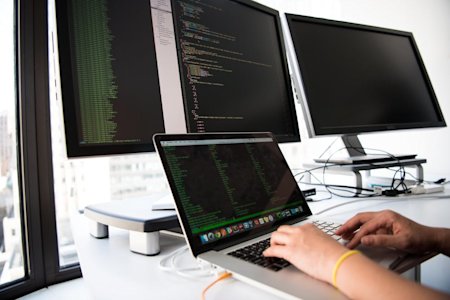 Hands typing on a laptop with two external monitors displaying code.