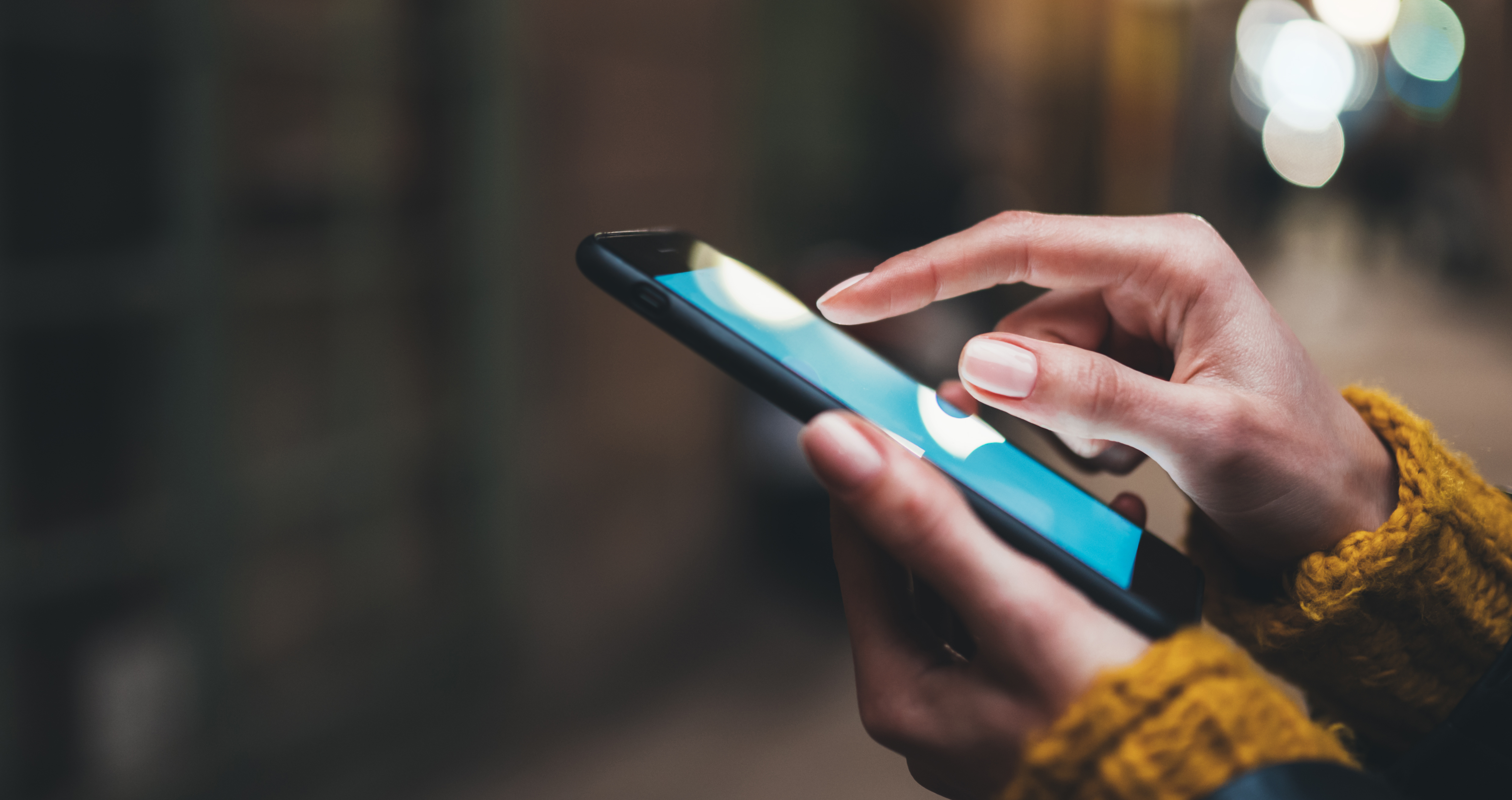 Close-up of a person’s hands using a smartphone outdoors. The person is tapping the screen with one finger and wearing a yellow knit sweater. The background is blurred with soft, warm lighting.