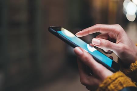 Close-up of a person’s hands using a smartphone outdoors. The person is tapping the screen with one finger and wearing a yellow knit sweater. The background is blurred with soft, warm lighting.