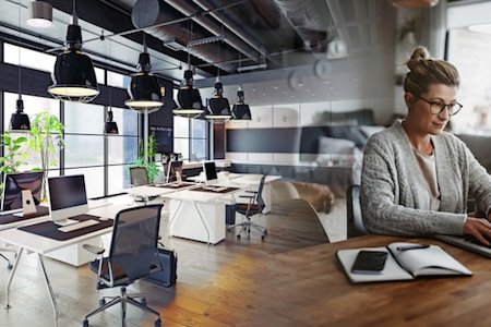 Woman is sitting on a gray couch in a modern office space with a laptop on her lap and a notebook next to her