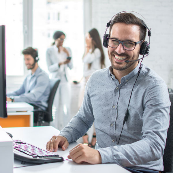 A smiling man wearing glasses and a headset sits at a desk with a computer in a bright office. In the background, two people work at computers and two others stand talking.