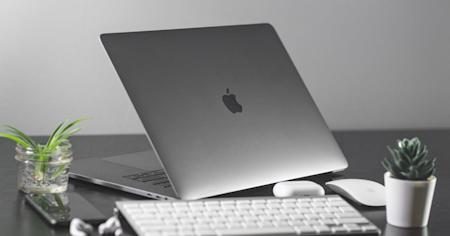 A Mac laptop, keyboard, mouse, and other items sitting on an office desk.
