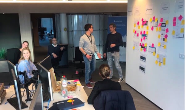 Five people are collaborating in a modern office. Two stand talking by a whiteboard covered in sticky notes, while three others work at desks with computers. The atmosphere appears casual and productive.