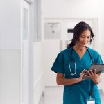 A healthcare professional in teal scrubs and a stethoscope stands in a hallway, smiling while using a digital tablet.