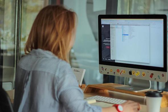 A person with light brown hair sits at a desk using a desktop computer with a screen displaying code or text files. A coffee cup, notebook, and other items are on the desk in a modern office setting.