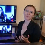 A woman sits indoors holding a remote, with a TV and a laptop behind her both displaying the same image of herself during a video call or recording. A plant is visible in the background.