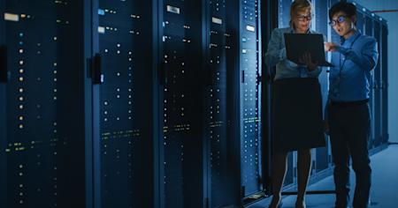 Two IT professionals reviewing data on a laptop in a server room filled with tall racks of network equipment.