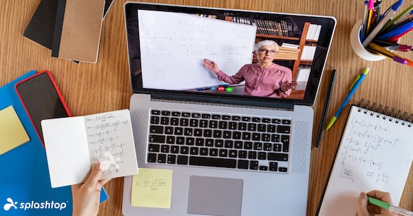 A person attends an online math class on a laptop, taking notes by hand while a teacher explains equations on a whiteboard during a video call. Notebooks, pens, and stationery are scattered on the wooden desk.