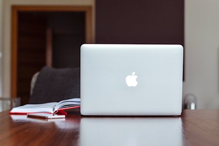 A Mac laptop on a desk next to a notebook.