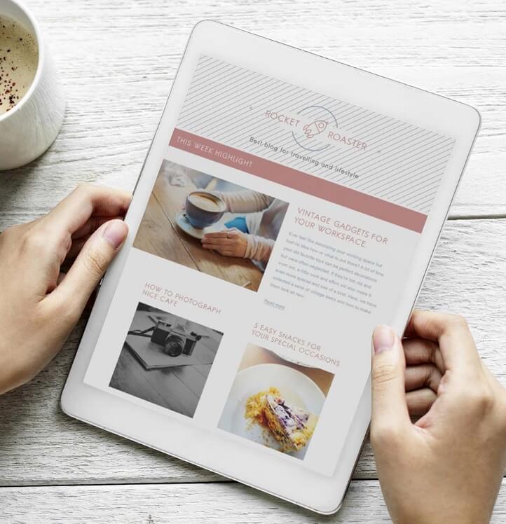 A person holds a white tablet displaying a digital magazine or blog featuring articles, a coffee cup photo, a camera image, and a dessert. A cup of coffee sits on the table beside the tablet.