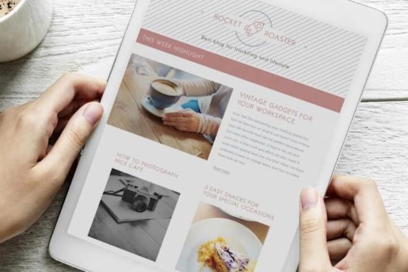 A person holds a white tablet displaying a digital magazine or blog featuring articles, a coffee cup photo, a camera image, and a dessert. A cup of coffee sits on the table beside the tablet.