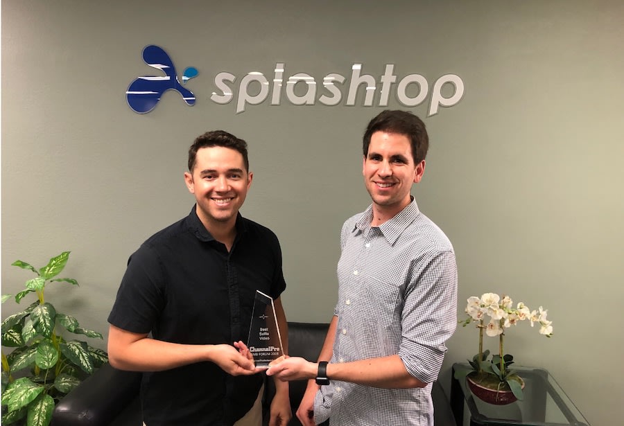 Two men stand smiling in front of a wall with the Splashtop logo. One holds a clear award. There are plants and a small table with orchids in the background.