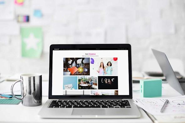 A laptop on a white desk displays a creative inspiration website. Next to it are a silver mug, a smartphone, a blue box, papers, and office supplies. The background shows a blurred wall with notes and photos.