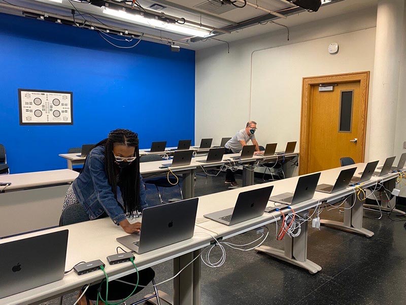 Two people work on separate laptops in a classroom with a blue accent wall. Many laptops are lined up on long tables, each connected with cables. The room appears to be used for tech setup or testing.