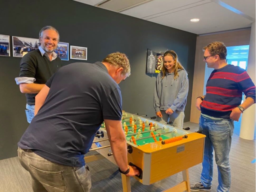 Four people are gathered around a foosball table, smiling and playing together in a casual indoor office space with photos on the dark wall behind them.